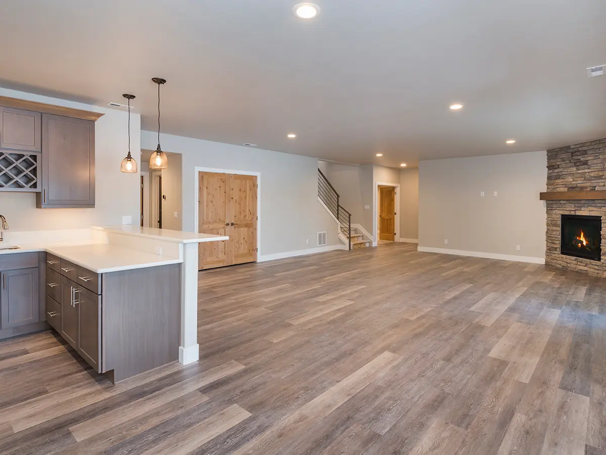 finished basement renovated white gray walls spotlights, wooden floor, fireplace, open-space kitchen with brown wood cabinets with white marble countertop